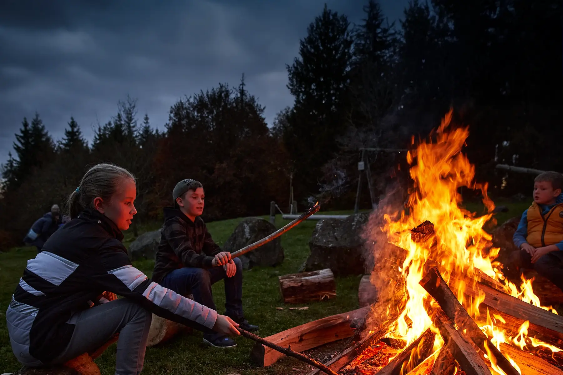 Kinder sitzen am Lagerfeuer und grillen Stockbrot bei Abenddämmerung